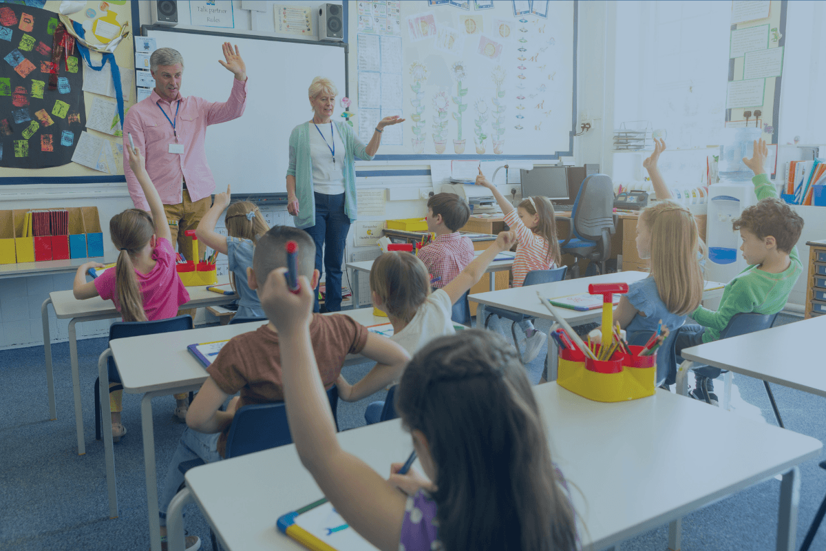 A few children are in class, raising hands, with 2 teachers in front of the board