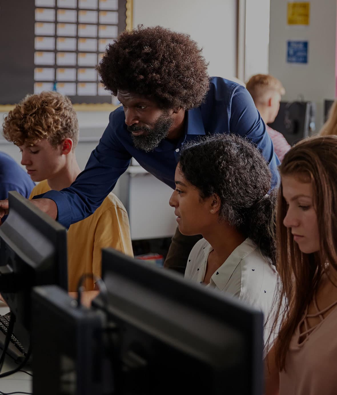 A teacher points at a computer screen, with students in the background